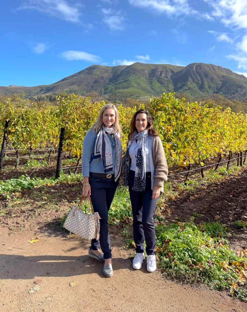 About Bernie and Petra of Travel Buddies Lifestyle. Two best friends standing in the vineyard of Groot Constantia with majestic mountains in the backdrop.